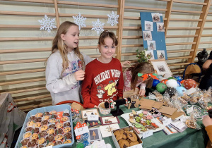 Uczennice stoją przy przygotowanym stoisku ze smakołykami i ozdobami świątecznymi
