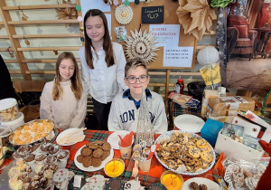 Uczennice siedzą (jedna stoi) przy przygotowanym stoisku ze smakołykami i ozdobami świątecznymi