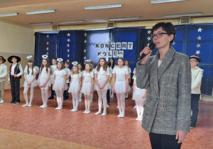 Pani Dyrektor przemawia podczas koncertu. W tle osoby występujące w imprezie.